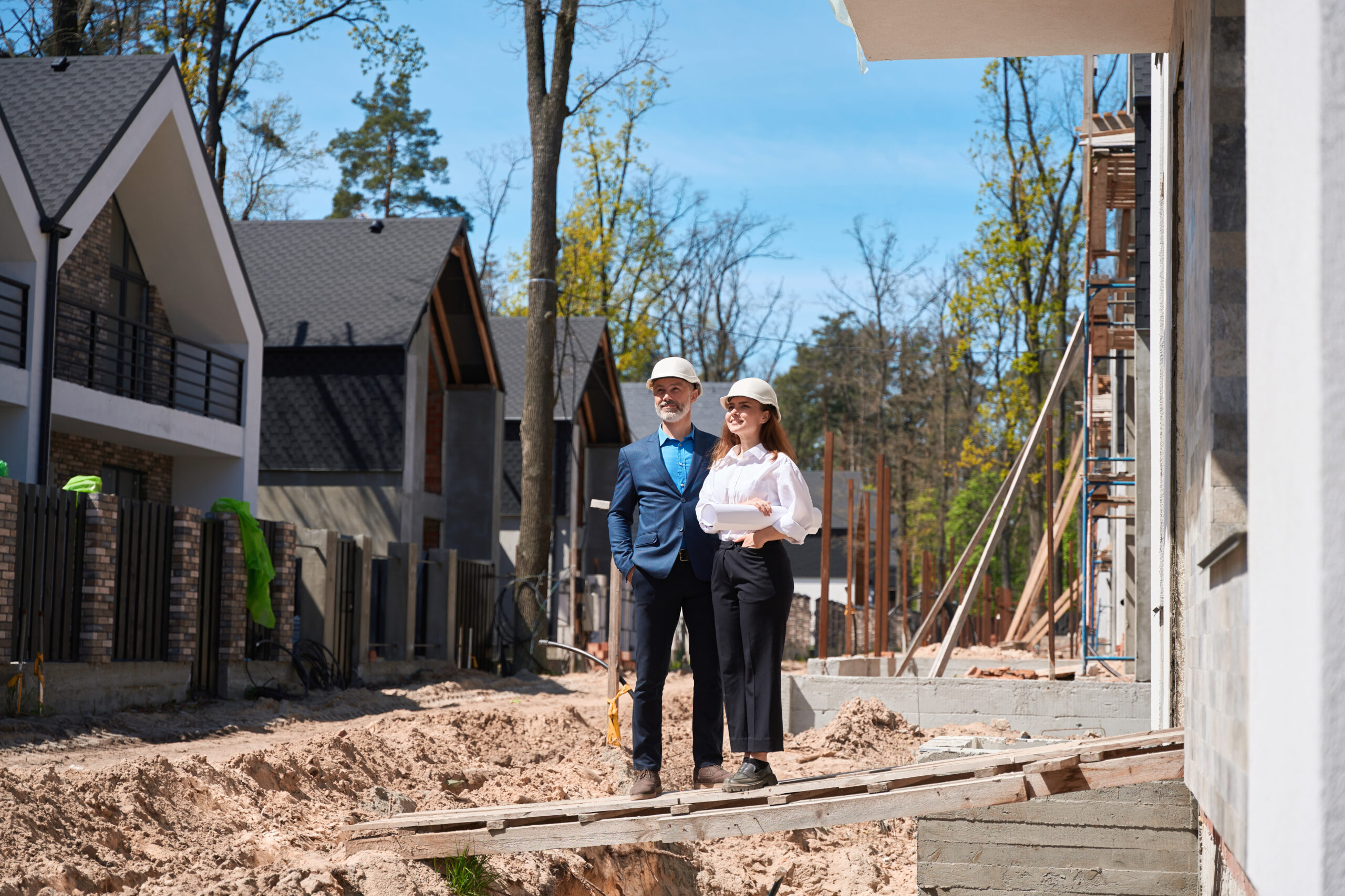 Two people in construction helmets stand on a wooden plank at a building site, smiling. They are surrounded by partially built homes and trees, under a clear blue sky.