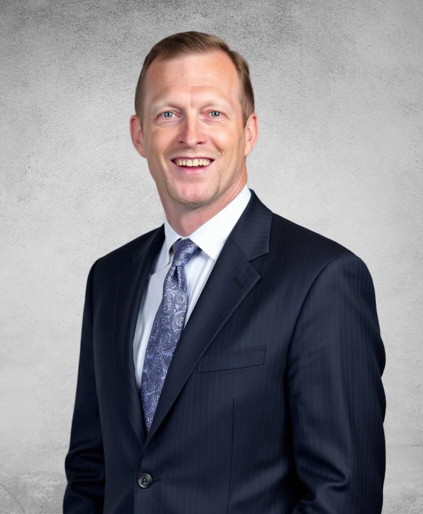 John Barlow, Senior Business Loan Consultant, smiles confidently in a dark suit and patterned tie. The background is a plain, textured gray surface.