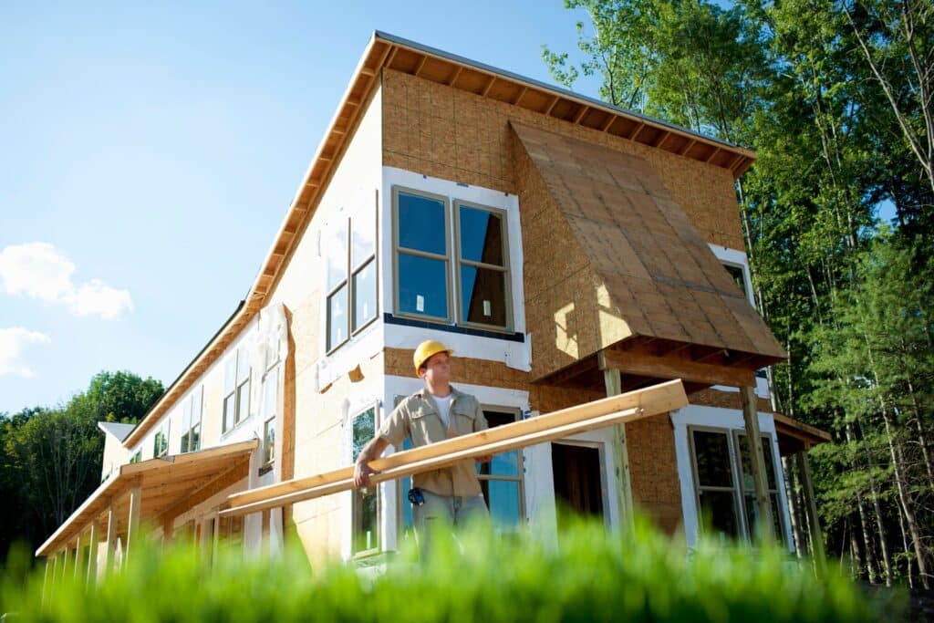 Construction worker carrying long lumber boards in front of a partially completed two-story home under construction on a sunny day.
