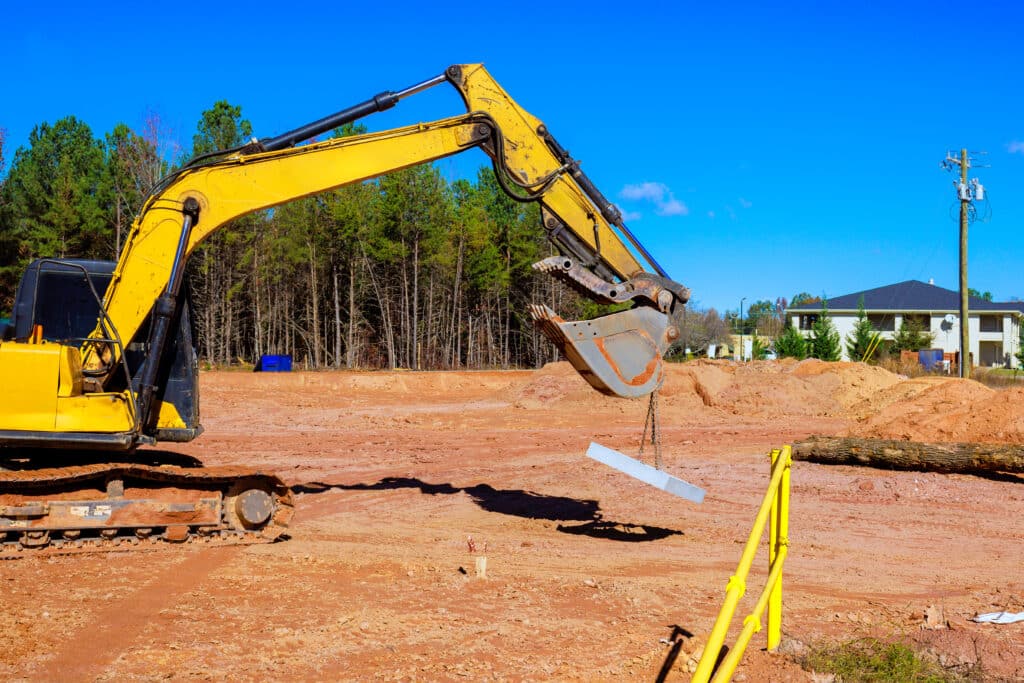 Yellow excavator lifting a concrete beam on a residential construction site with cleared land and framing in progress