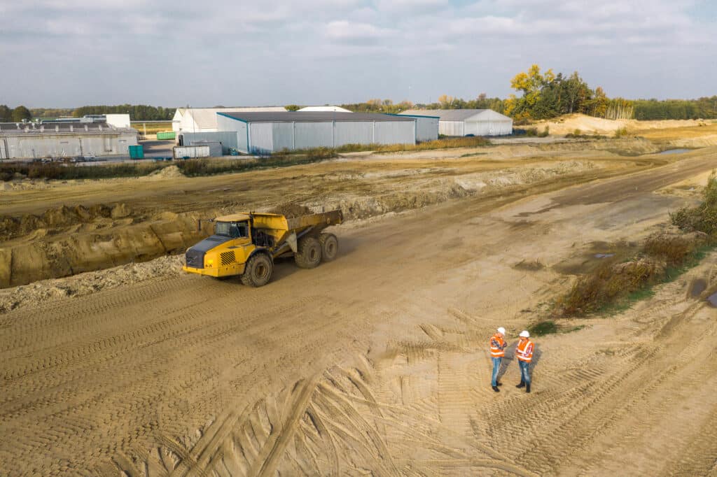 Construction site with a large yellow dump truck carrying soil and two workers in safety vests and hard hats reviewing plans on the dirt ground, with industrial buildings and trees in the background.